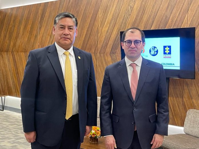 El Fiscal General Francisco Barbosa junto al Fiscal General de Bolivia están parados uno al lado del otro. Al fondo se ve un televisor con los logos de las Fiscalías.
