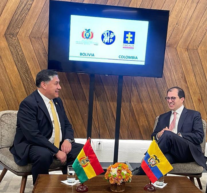 El Fiscal General de Colombia, Francisco Barbosa, está sentado en una sala y al frente suyo está su par de Bolivia, Juan Lanchipa. Sobre una mesa se aprecian las banderas de ambos países.