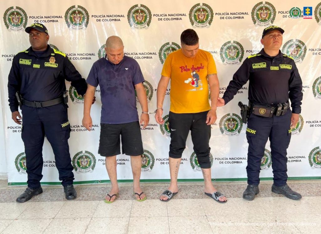 Los capturados se encuentran de frente y custodiados por dos uniformados de la Policía Nacional. Detrás se observa un backing de la Policía Nacional.
