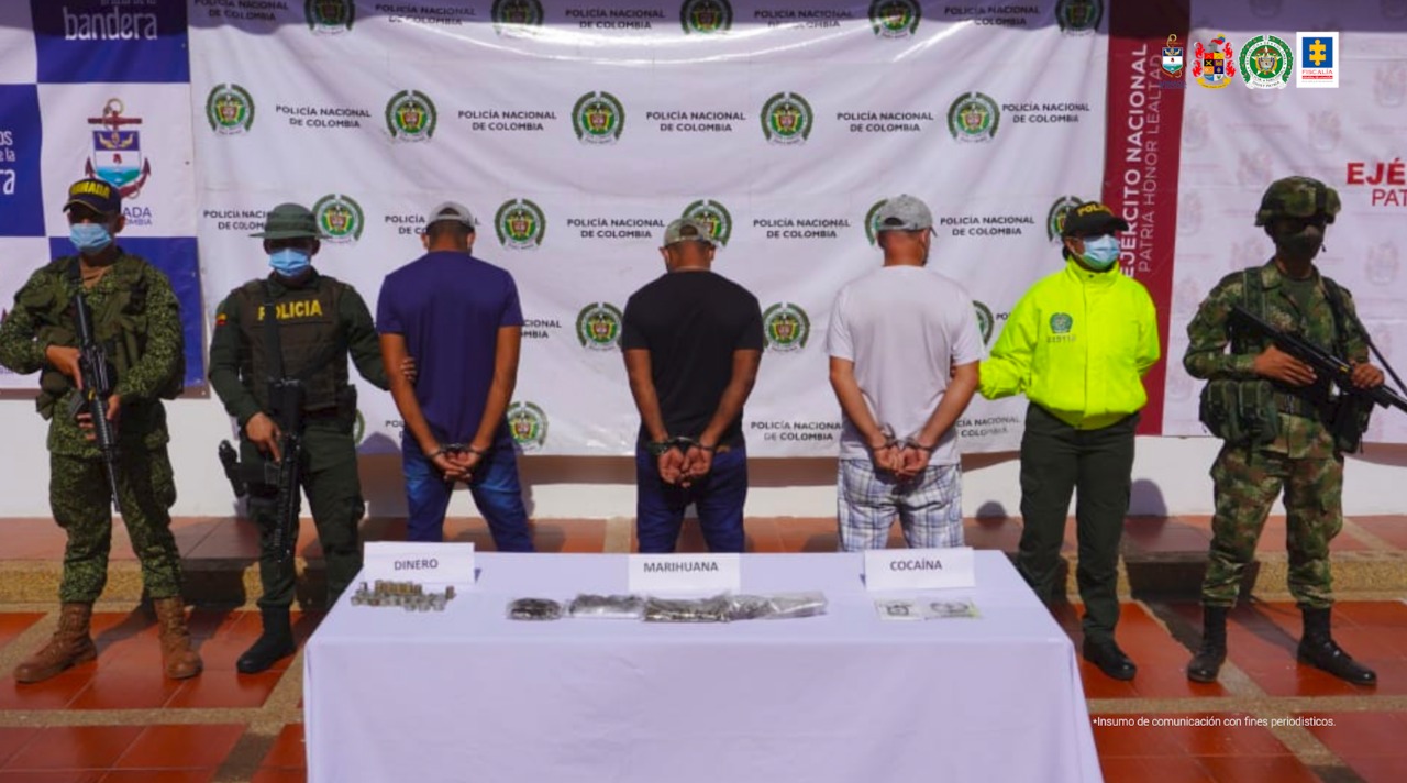 Los seis capturados están esposados a su espalda, y en una mesa están algunos de los elementos incautados y están custodiados por hombres de la Policía nacional, detrás hay un pendón institucional.