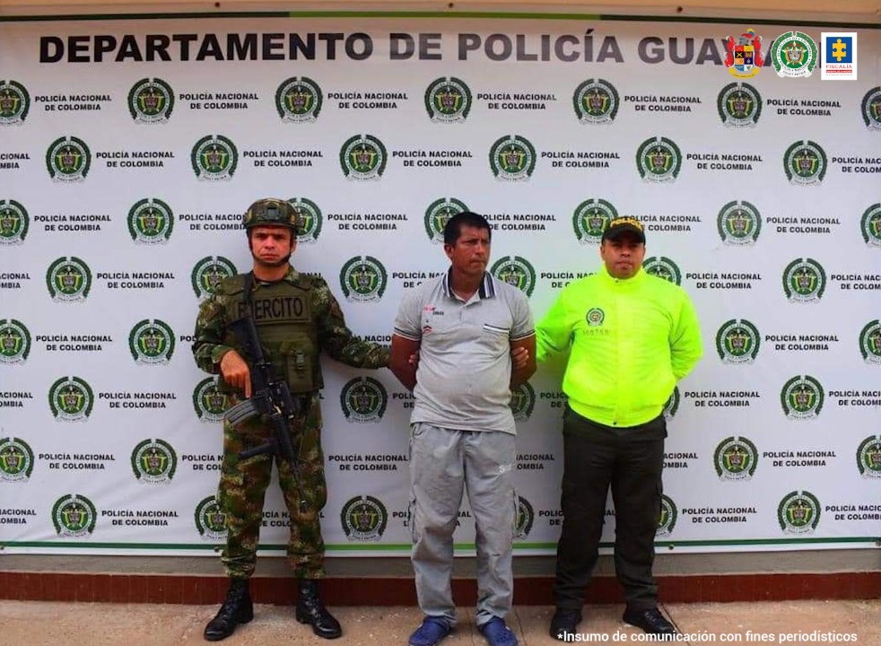 En la fotografía se ven tres personas, una de ellas el capturado, las otras dos personas son integrantes de la Policía y el Ejército Nacional. En la parte posterior se ve un baking de la Policía Nacional con un letrero que indica Departamento de la Policía de Guaviare. 