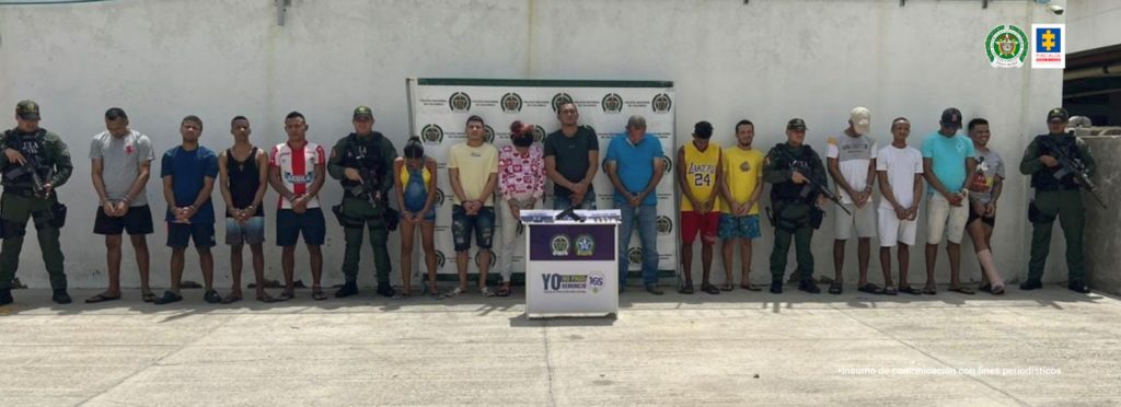 En la imagen aparecen 14 hombres y dos mujeres de pie, con los brazos hacia adelante esposados, custodiados por cuatro agentes de la Policía Nacional con una mesa en la que se encuentran unas armas y municiones incautadas. Detrás de ellos hay un pendón de esa institución.