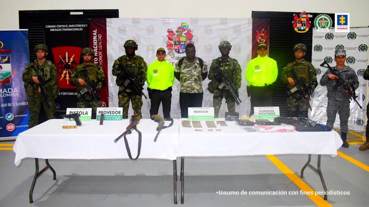 En la fotografía se observan nueve personas, una de ellas es el capturado, seis de ellas uniformados del Ejército Nacional y uno uniformado de la Policía Nacional. En la parte delantera hay dos mesas donde se encuentra material incautado como armas, municiones y computadores. En la parte de atrás se encuentran cuatro backings: tres del Ejército y uno de la Policía.