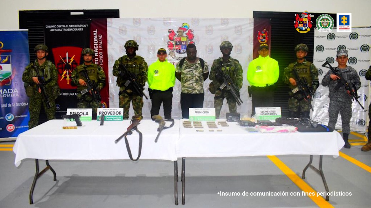  En la fotografía se observan nueve personas, una de ellas es el capturado, seis de ellas uniformados del Ejército Nacional y uno uniformado de la Policía Nacional. En la parte delantera hay dos mesas donde se encuentra material incautado como armas, municiones y computadores. En la parte de atrás se encuentran cuatro backings: tres del Ejército y uno de la Policía. 