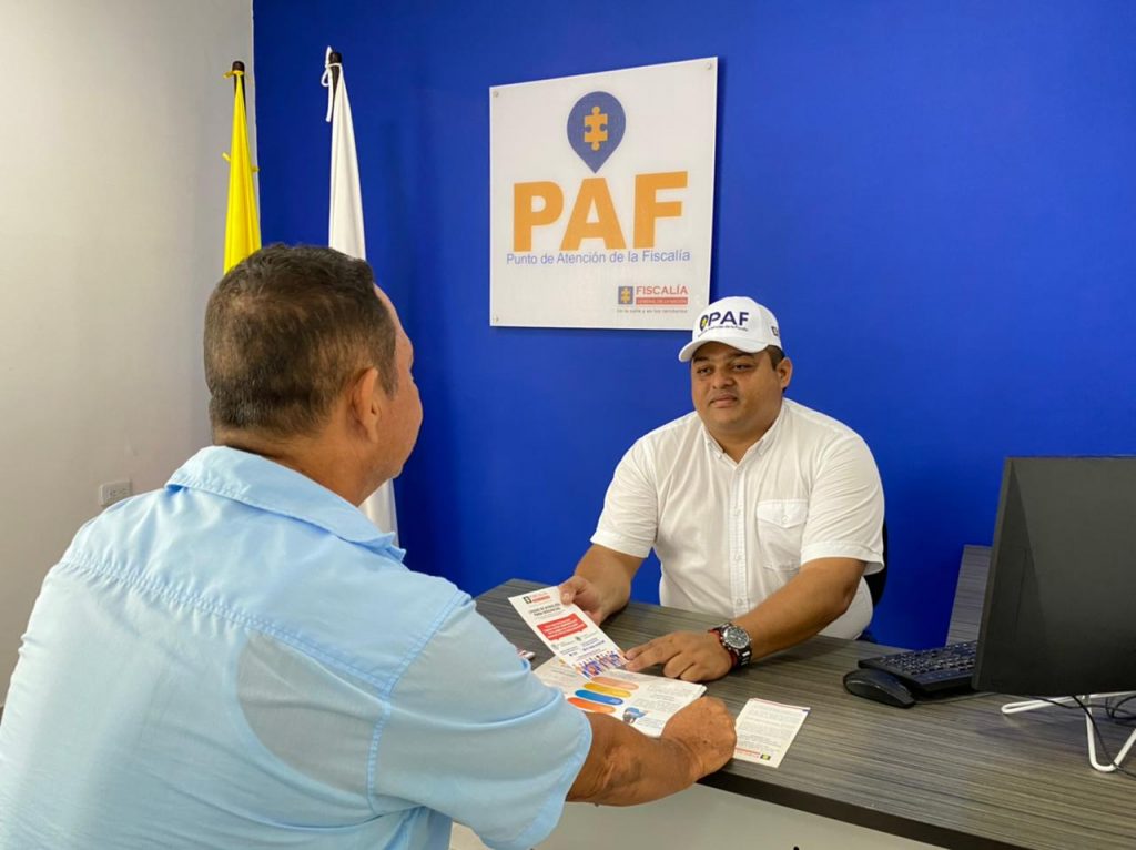 En la imagen se ve un funcionario de la Fiscalía atendiendo a un ciudadano, en un escritorio.