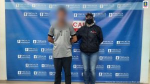 En la fotografía aparece un hombre con el rostro difuso vestido de camiseta gris con jean negro, custodiado por un uniformado del CTI. Detrás de ellos hay un pendón azul que dice Fiscalía General de la Nacional.