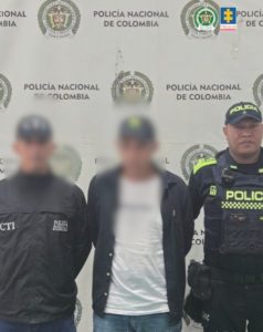 En la fotografía aparece un hombre de pie, de frente, con los brazos atrás esposados, tiene el rostro difuso, está vestido con una gorra negra, camiseta blanca, saco negro y un jean azul. Se encuentra custodiado por un uniformado del CTI y otro de la Policía Nacional. 
