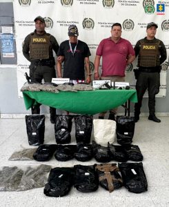 En la imagen están dos hombres morenos, de pie, de frente, con los brazos esposados, uno se encuentra vestido con camiseta negra y el otro con camiseta roja. Están custodiados por uniformados de la Policía Nacional. Delante de ellos hay una mesa con el material bélico y la droga incautada. 