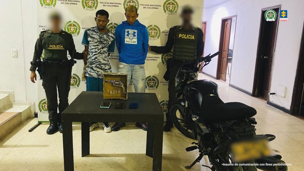 En la imagen están dos hombres morenos, delgados de pie,con los brazos atrás esposados, custodiados por dos uniformados de la Policía Nacional.