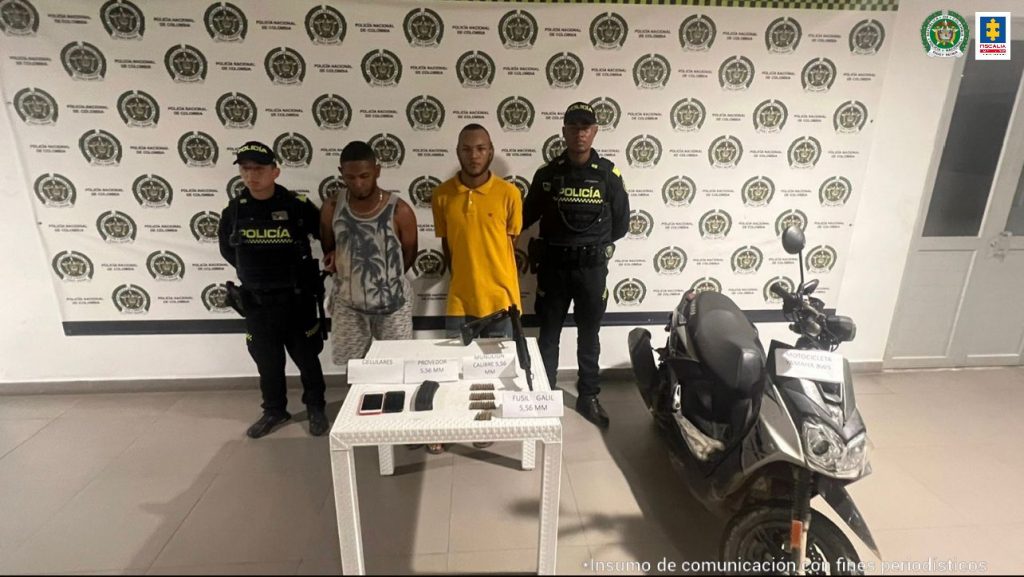 En la fotografía aparecen dos hombres morenos, uno vestido de camiseta amarilla con jean y el otro de camisilla gris con bermuda estampada, custodiados por dos agentes de la Policía Nacional. Detrás de ellos hay un pendón de esa institución.