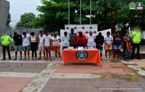 En la imagen aparece un grupo de personas de pie, con los brazos atrás, entre los que se encuentran hombres y mujeres, con las cabezas agachadas, custodiados por dos agentes de la Policía Nacional, delante de ellos hay una mesa con la droga incautada. 