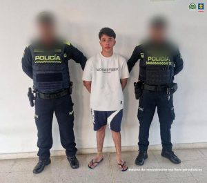 En la fotografía está un hombre joven, de pie, de frente, blanco, con los brazos atrás esposado, cabellos lisos negros, vestido con una camiseta blanca, bermuda y sandalias negras. Se encuentra custodiado por dos uniformados de la Policía Nacional. 