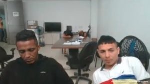 En la imagen están dos hombres, uno de tez morena vestido de buzo negro y el otro de color blanco vestido con camiseta blanca. Ambos se encuentran sentadas en una oficina donde asisten de manera virtual a la audiencia que los procesó.