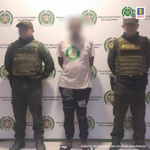 En la fotografía hay un hombre de pie con el rostro difuso, vestido de camiseta blanca con sudadera negra y sandalias negras, custodiado por dos uniformados de la Policía Nacional. Detrás de ellos hay un pendón de esa institución.