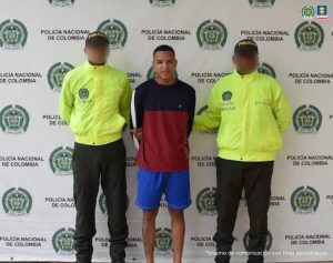 En la fotografía aparece un hombre de pie, delgado, con los brazos atrás, moreno, de cabellos cortos crespos negros, vestido con un buzo rojo, bermuda azul. Se encuentra custodiado por dos uniformados de la Policía Nacional.