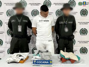En la fotografía aparece un hombre de pie, con los brazos atrás, delgado, alto, trigueño, de cabellos negros, vestido con camiseta y pantalón blanco, custodiado por dos agentes de la Policía Nacional. Delante de ellos hay una mesa con la ropa incautada.