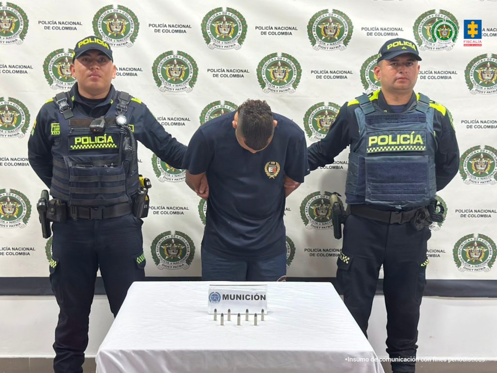 En la fotografía está un hombre de baja estatura de pie, de frente, con la cabeza agachada, cabellos negros, vestido con camiseta oscura y está custodiado por dos uniformados de la Policía Nacional. Delante de ellos hay una mesa con las municiones incautadas. 