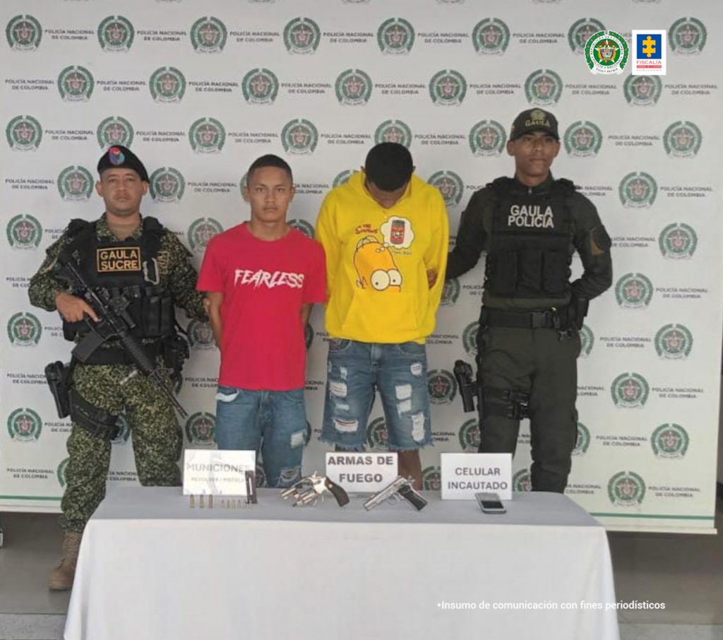 En la imagen aparecen dos hombres de pie, uno con la cabeza agachada vestido de buzo amarillo y jean azul y el otro de camiseta roja con bermuda de jean azul, custodiado por dos agentes de la Policía Nacional. Delante de ellos hay una mesa con las armas incautadas.