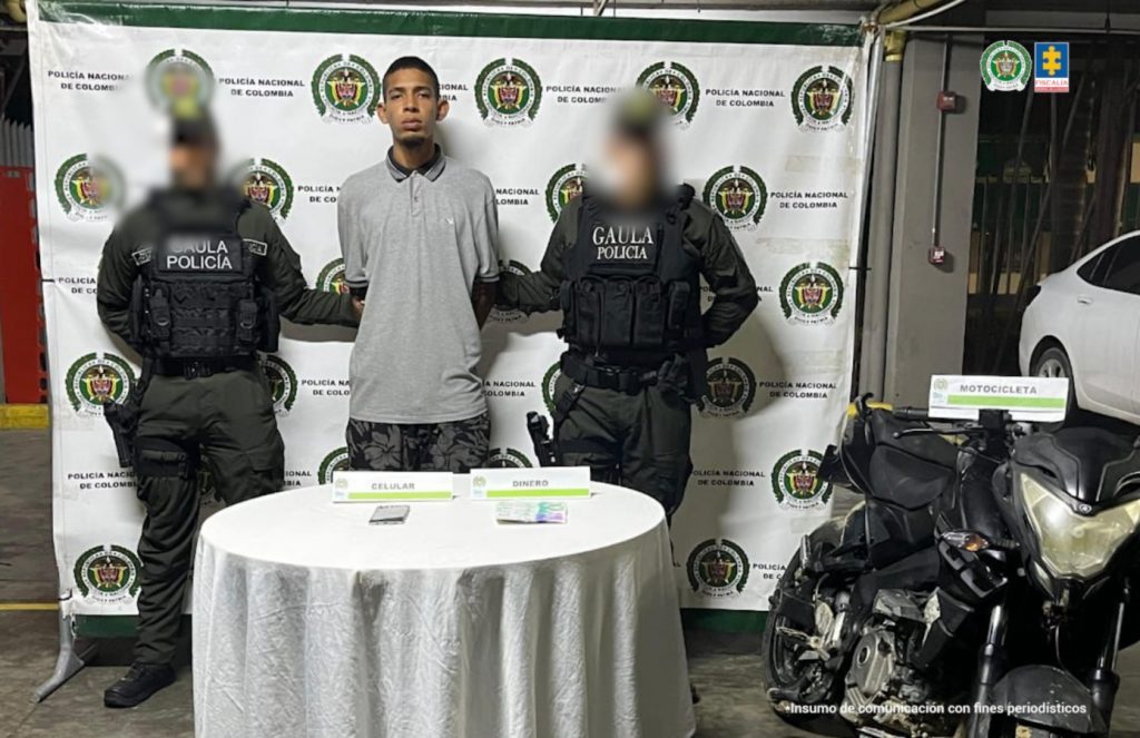 En la fotografía está un hombre joven, delgado, de pie, moreno, cabellos negros, con los brazos atrás esposados, vestido con camiseta gris, pantalón negro estampado y custodiado por dos uniformados de la Policía Nacional.