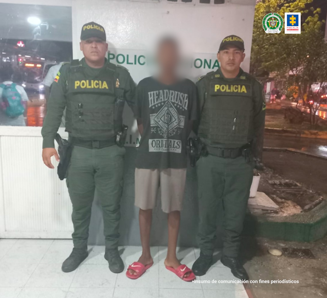 En la fotografía aparece un hombre con camiseta negra, bermuda beis y chanclas rojas, custodiado por dos agentes de la Policía Nacional delante de una pared blanca en la cual se lee Policía Nacional.