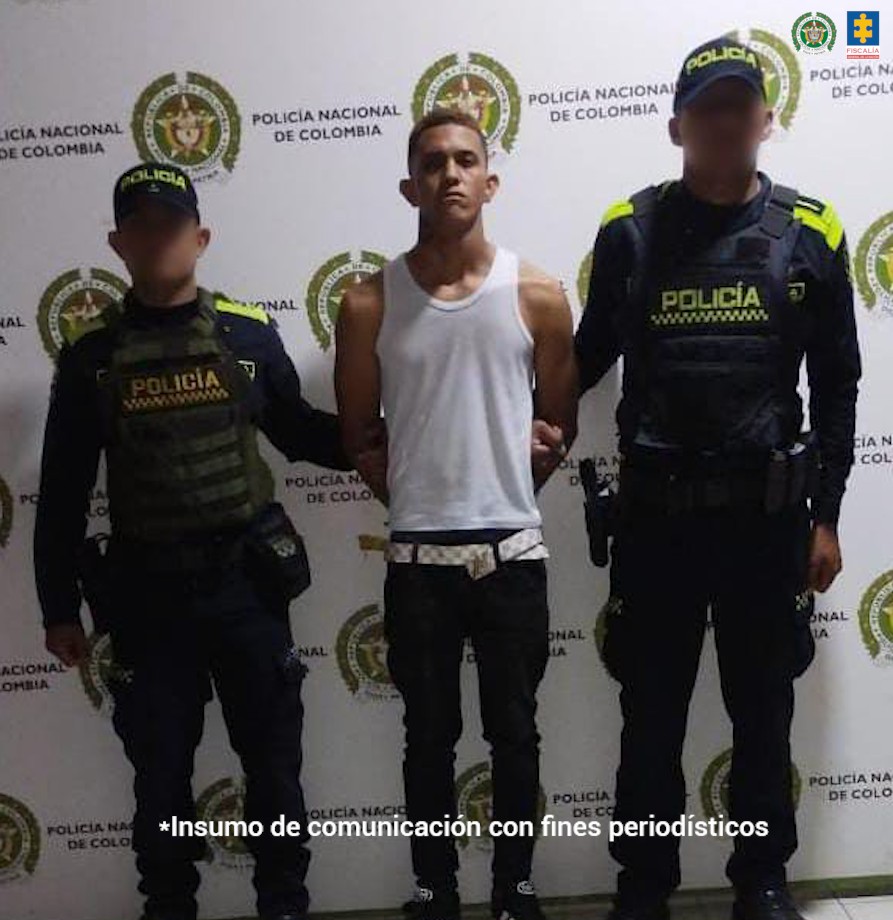 En la imagen está un hombre joven de pie, delgado, de frente, trigueño, con los brazos atrás esposados, vestido con una camisilla y correa blanca, pantalón y tenis negro y se encuentra custodiado por dos uniformados de la Policía Nacional.