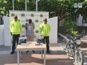 En la imagen se observa a un hombre de camiseta gris con pantalón beis, custodiado por dos agentes de la Policía Nacional, delante de un pendón de esa institución. También se divisa una mesa blanca con el arma y el estupefaciente que le fue incautado.