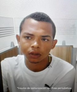 En la foto está el rostro de un hombre joven, moreno, cabellos cortos crespos, de frente ancha amplia, labios gruesos, vestido con una camiseta blanca. Se encuentra sentado en una silla al interior de una oficina asistiendo virtualmente a la audiencia donde se le procesó.