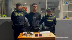 En la fotografía está un hombre joven de pie, de frente, moreno, cabellos negros lisos cortos, orejas grandes, se encuentra vestido con una camiseta y un jean negro y está custodiado por dos uniformados de la Policía Nacional en las afueras de un CAI.