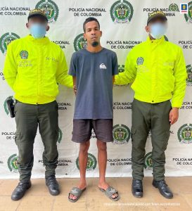 En la fotografía hay un hombre delgado vestido de camiseta azul oscura con bermuda negra y sandalias grises. Se encuentra acompañado por dos uniformados de la Policía Nacional. Detrás de ellos está un pendón de esa institución.