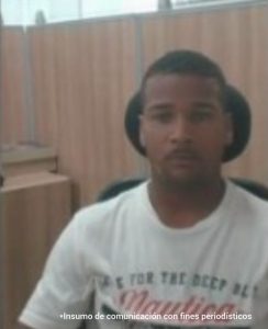 En la fotografía aparece el rostro de un hombre joven, moreno con barba, cabello corto rizado, vestido de camiseta blanca con letras negras y rojas, sentado en una silla negra escuchando la audiencia donde le fueron imputados los delitos por la Fiscalía. 