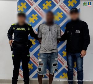 En la imagen se encuentra un hombre moreno de pie, con los brazos atrás, el rostro difuminado, vestido con una camiseta gris y una bermuda azul, custodiado por un uniformado del CTI y otro de la Policía Nacional. Detrás de ellos hay un pendón de la Fiscalía.