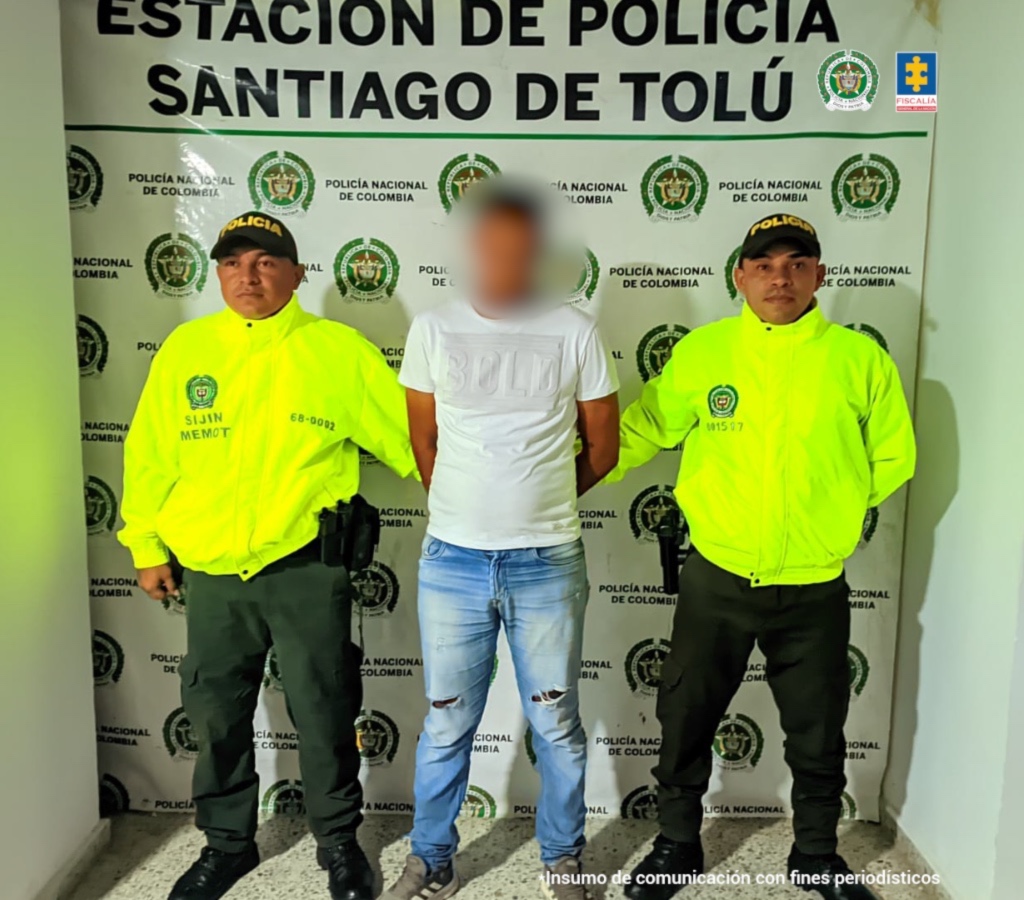 En la fotografía aparece un hombre de pie, con el rostro difuso, con los brazos atrás esposados, de contextura mediana, vestido con camiseta blanca, jean azul y tenis negro. Se encuentra custodiado por dos uniformados de la Policía Nacional.