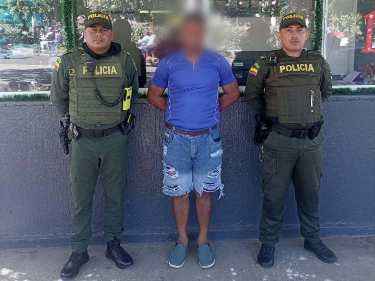 En la imagen se observa a un hombre con rostro difuso, camiseta azul y bermuda de jean, custodiado por dos agentes de la Policía Nacional.