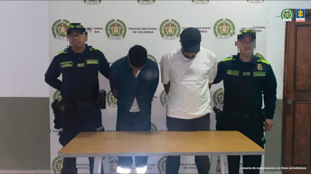 En la fotografía están dos hombres de pie, de frente, con las cabezas agachadas ambos, uno es de baja estatura y se encuentra vestido con camisa y pantalón azul oscuro, el otro es alto, tiene una gorra negra en la cabeza y está vestido con camiseta blanca y pantalón negro. Se encuentran custodiado por dos uniformados de la Policía Nacional.