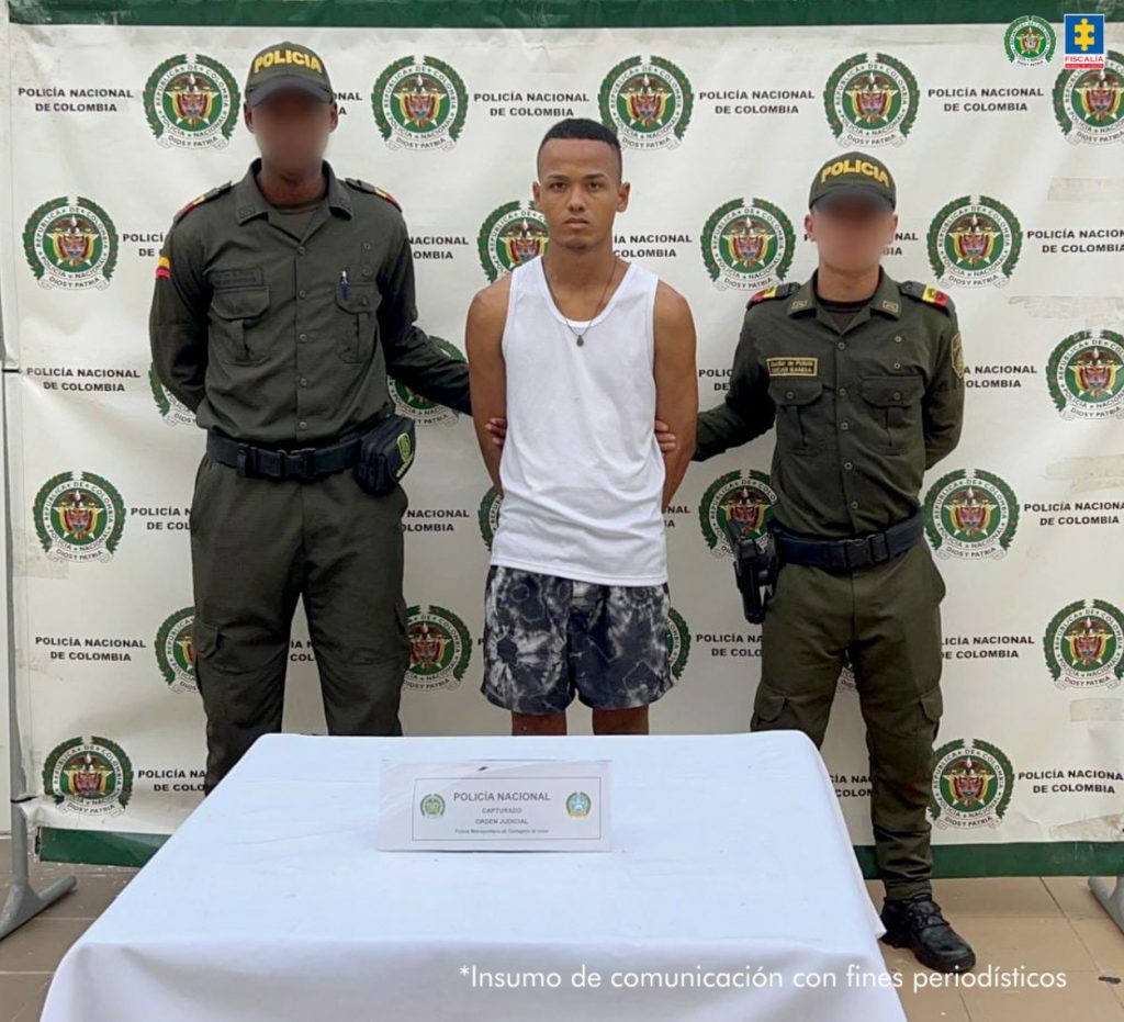 En la fotografía aparece un hombre joven delgado de pie, moreno, con los brazos atrás esposado, cabellos negros crespos cortos, vestido con camisilla blanca y bermuda estampada negra con blanca. Se encuentra custodiado por dos uniformados de la Policía Nacional.