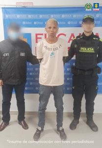 En la fotografía está un hombre delgado de pie, de frente, con los brazos atrás esposados, con los cabellos tinturados de amarillo, vestido con una camiseta blanca, jean azul y zapatos negros. Se encuentra custodiado por un uniformado del Cuerpo Técnico de Investigación y otro de la Policía Nacional. Detrás de ellos hay un pendón azul de la Fiscalía.