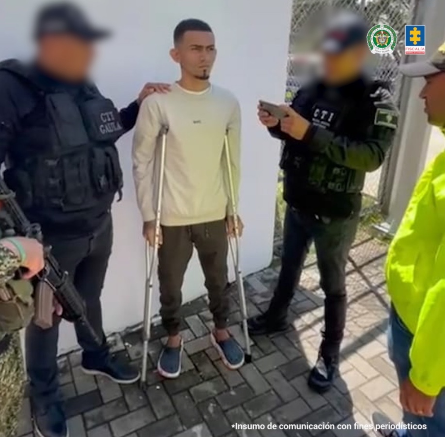 En la foto se encuentra un hombre de pie, trigueño, joven, tiene muletas, vestido con buzo beige y pantalón café, sandalias azules, custodiado por uniformados del CTI y la Policía Nacional.