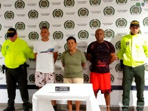 En la imagen aparecen dos hombres y una mujer custodiados por dos agentes de la Policía Nacional. Los hombres están vestidos uno con camiseta blanca y bermuda estampada y el otro con camiseta negra y bermuda roja. La mujer viste una camiseta verde y una bermuda blanca.