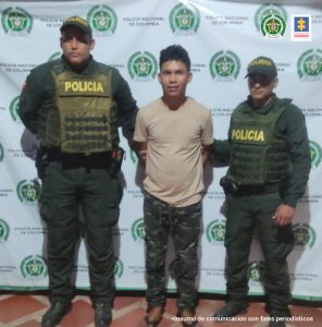 En la imagen hay un hombre de pie vestido de camiseta beis con pantalón estampado verde, tiene los brazos atrás y se encuentra custodiado por dos agentes de la Policía Nacional.