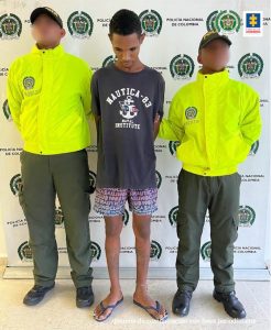 En la fotografía aparece un hombre delgado, moreno de pie, con los brazos atrás, tiene la cabeza agachada, vestido de camiseta negra, bermuda de color rosada con morada y sandalias azules. Se encuentra custodiado por dos agentes de la Policía Nacional.