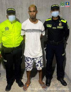 En la fotografía aparece un hombre de pie, alto, moreno, con los brazos atrás, de cabellos cortos, crespos, rubio teñido , con barba, vestido de camiseta blanca y bermuda azul con blanca, custodiado por dos agentes de la Policía Nacional.