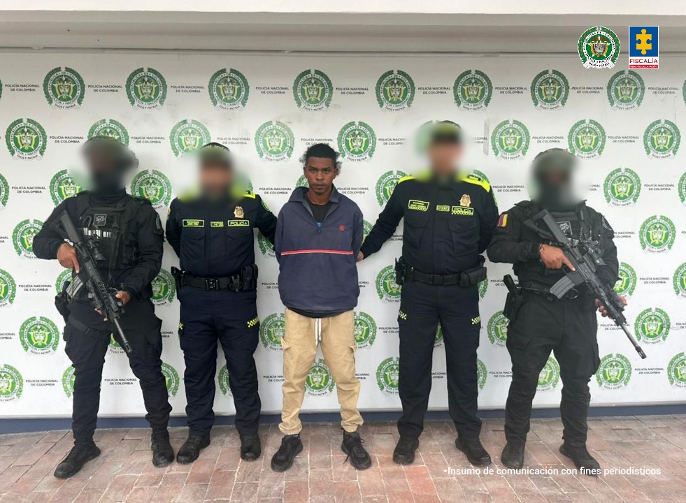 En la fotografía aparece un hombre joven, moreno, de pie, de frente, delgado, con los brazos atrás esposado, cabellos negros crespos, está vestido con un buzo azul y camiseta negra debajo, un pantalón beige y zapatos negros. Se encuentra custodiado por dos uniformados de la Policía Nacional.