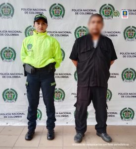 En la imagen se observa a un hombre de tez blanca vestido con pantalón, camisa y zapatos negros, custodiado por una uniformada de la Policía Nacional, delante de un pendón de esa institución.