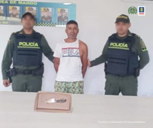 En la fotografía aparece un hombre de camiseta roja con bermuda rosada, sin zapatos, custodiado por dos agentes de la Policía Nacional. Detrás de ellos hay un pendón de esa institución.