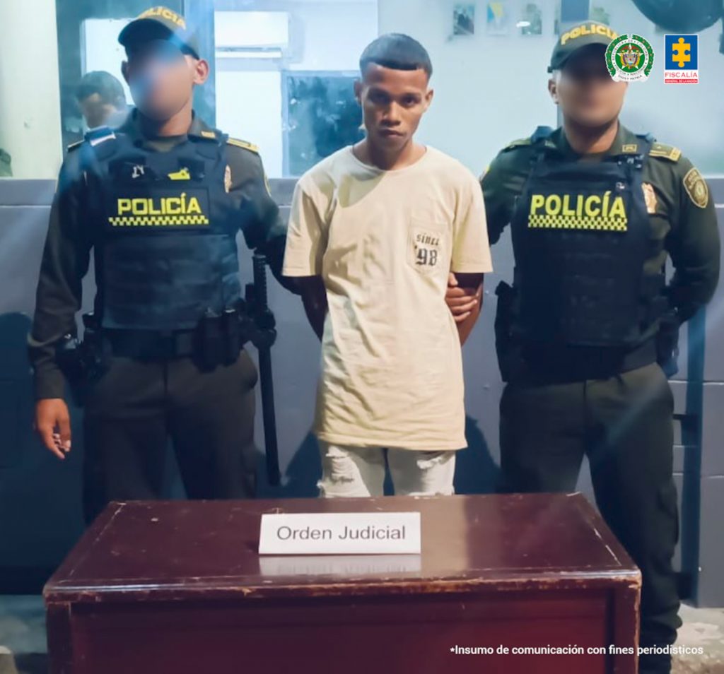 En la fotografía está un hombre de pie, con los brazos atrás esposados, delgado, moreno, joven, de orejas grandes, cabellos cortos crespos, vestido con una camiseta beige y un pantalón blanco. Se encuentra custodiado por dos uniformados de la Policía Nacional.