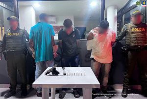 En la fotografía aparecen tres hombres de pie, esposados, con las cabezas agachadas, en un CAI de la Policía, custodiado por dos uniformados. Delante de ellos está una mesa con el arma y las municiones incautadas.