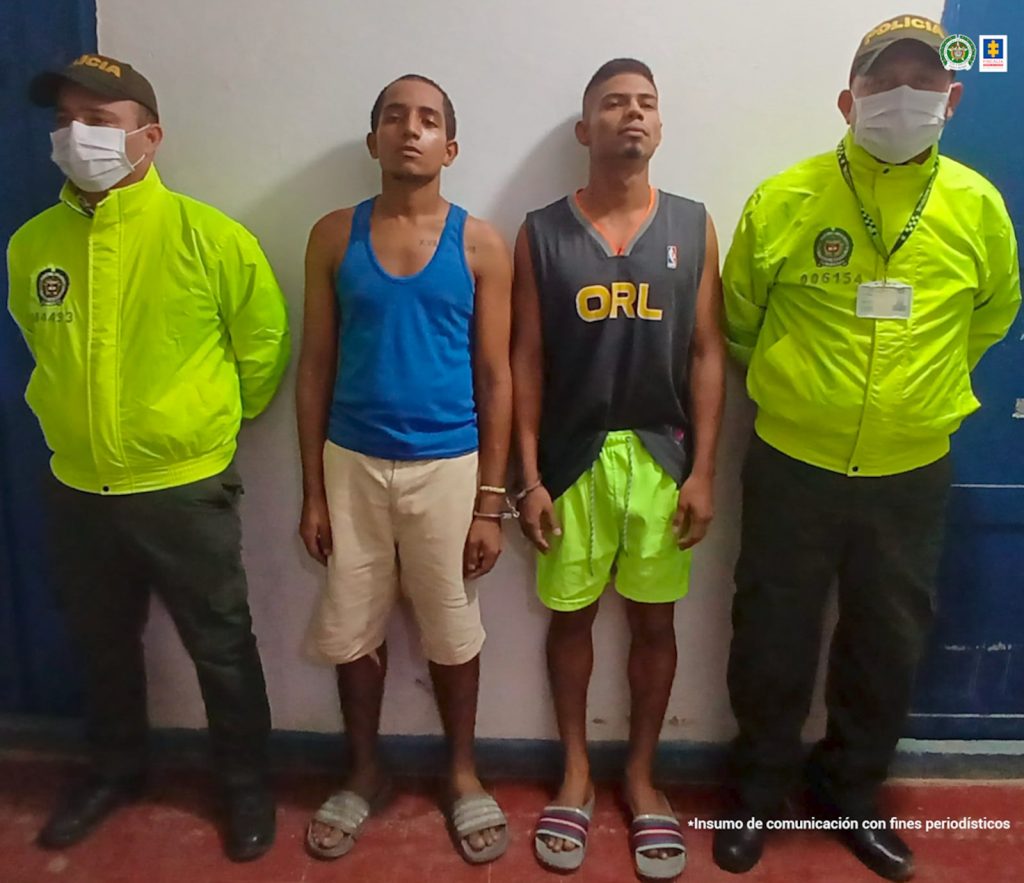En la imagen aparecen dos hombres delgados de pie, uno vestido de camisilla azul y bermuda blanca y el otro con camisilla gris y bermuda verde, custodiados por dos agentes de la Policía Nacional.