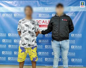 En la fotografía está un hombre de pie, con los brazos atrás, el rostro difuso, vestido de bermuda amarilla con azul y camiseta blanca con negra, custodiado por un uniformado del CTI. Detrás de ellos hay un pendón azul de la Fiscalía General de la Nación.