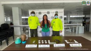 En la fotografía aparece una mujer morena, de contextura gruesa, cabello trenzado largo oscuro, vestida con blusa verde y un jean. Se encuentra custodiada por dos agentes de la Policía Nacional. Detrás de ella hay un pendón de esa institución y delante de ella una mesa con bolsas de estupefacientes y un arma de fuego.