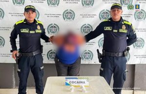 En la fotografía está una mujer de pie, de frente, agachada, con los brazos atrás esposados, vestida con una camiseta azul y un pantalón negro. Se encuentra custodiada por dos uniformados de la Policía Nacional.